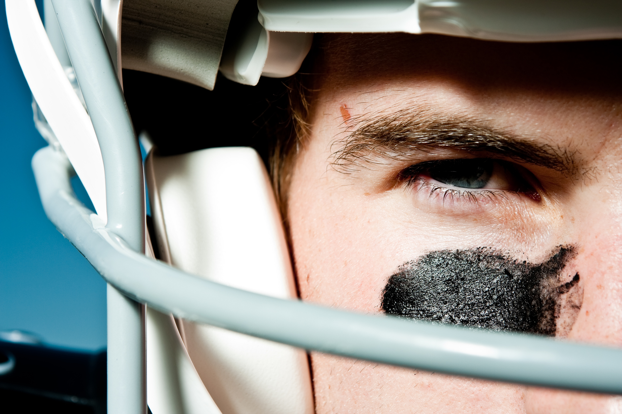 Young Man in his 20s wearing helmet and pads for American style football holding a football.  He has eye black on his face and has blue eyes.  He has a very serious and competitive look on his face.