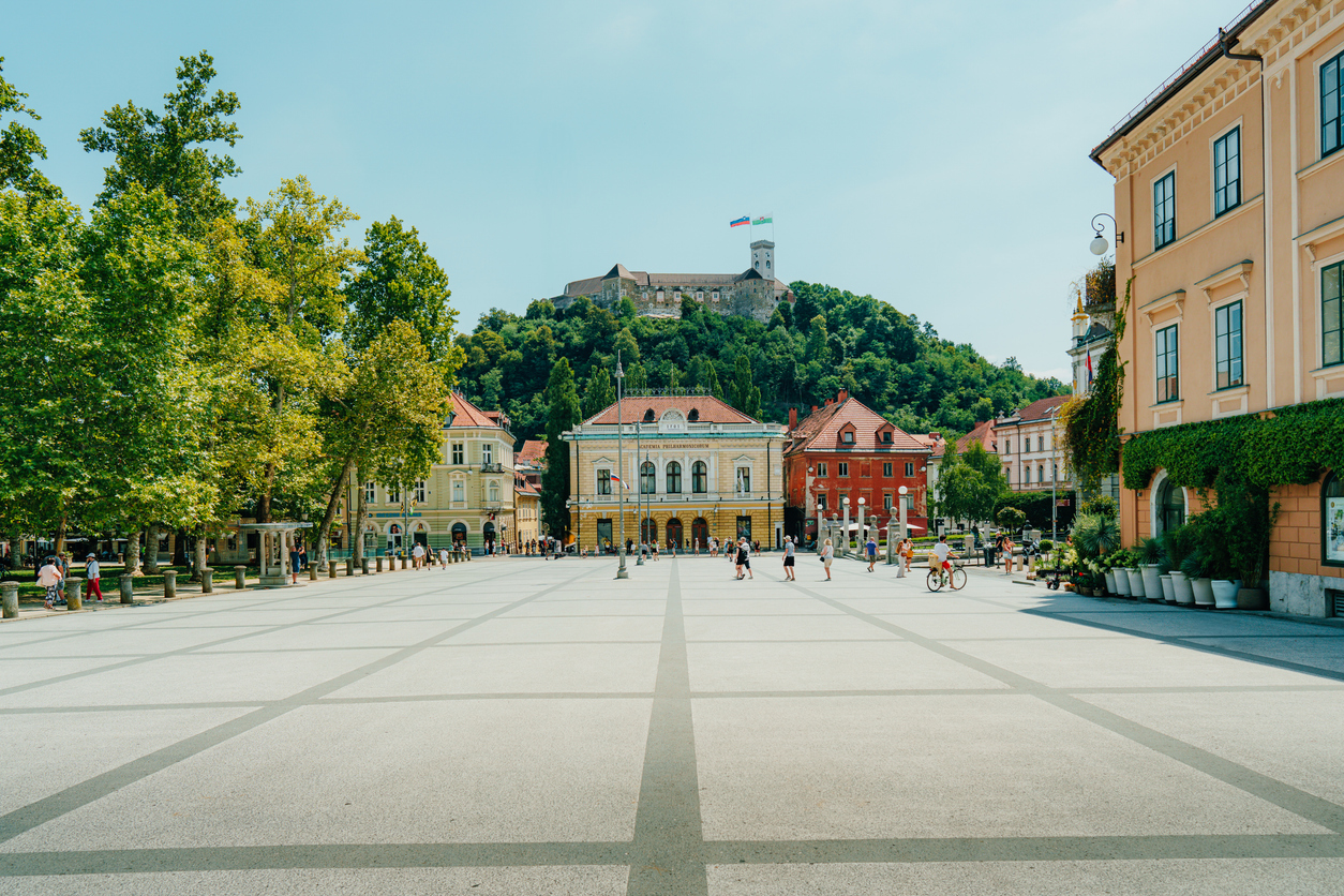 A vibrant city square in Ljubljana, Slovenia, with people enjoying a sunny day, historic buildings, and a castle in the background.